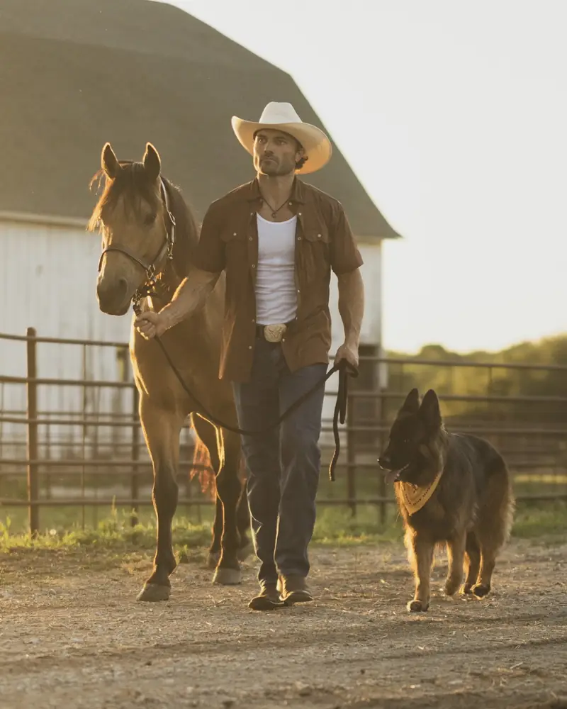 Steven leading a buckskin horse past the barn with his German Shepherd